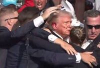 Trump yang berada di atas panggung terdiam,  Pengawal mengelilinginya.